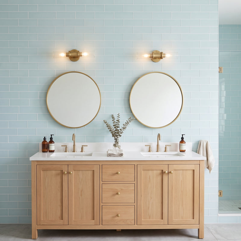 A double-sink bathroom vanity with a light oak wood cabinet, white countertop, and two round gold-framed mirrors mounted on a pale blue subway tile wall. Two brass wall sconces are above the mirrors, and small amber bottles and a vase with dried stems sit on the counter.