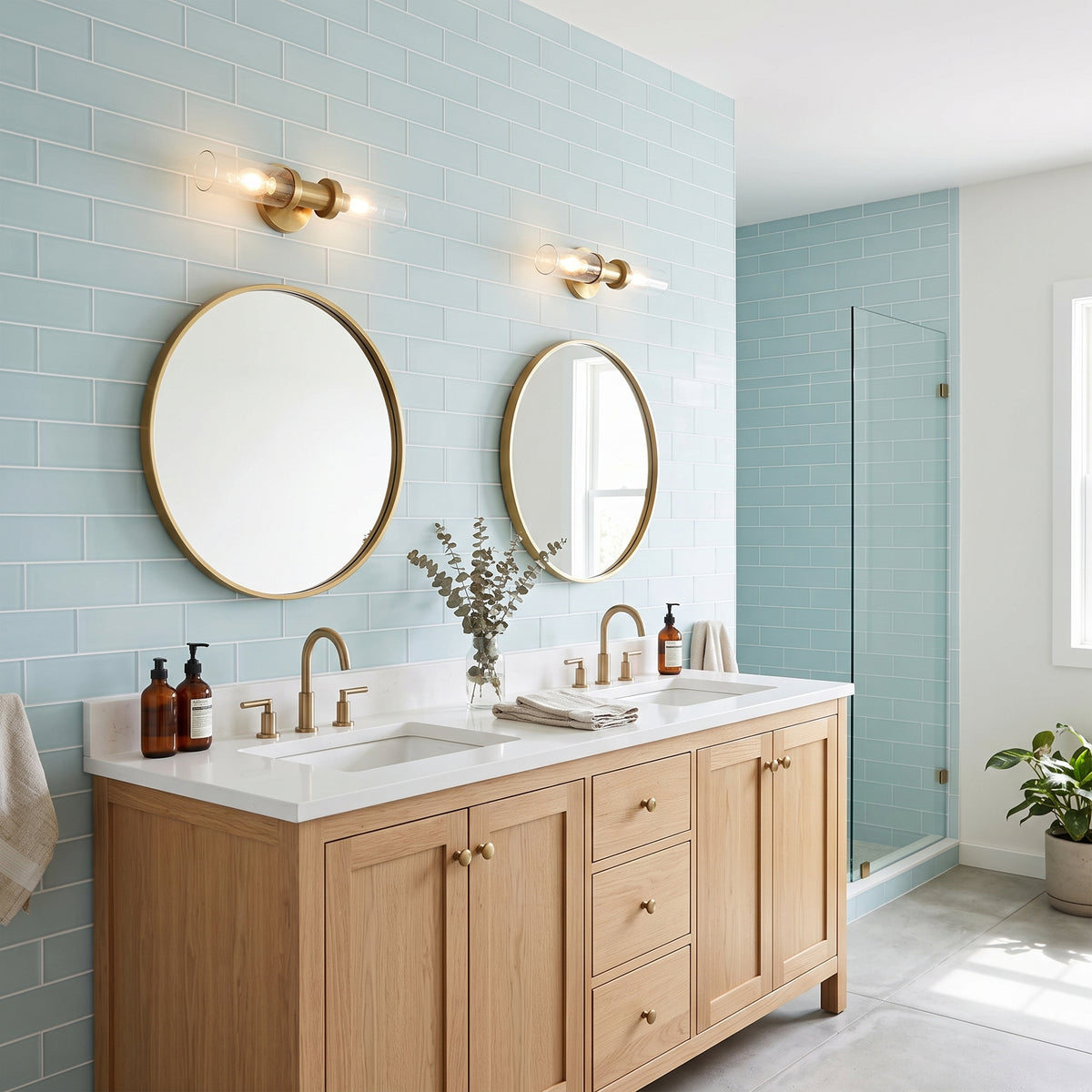 A modern double-sink bathroom vanity with a light wood cabinet, white countertop, round gold-framed mirrors, and brass fixtures, set against pale blue subway tile walls.
