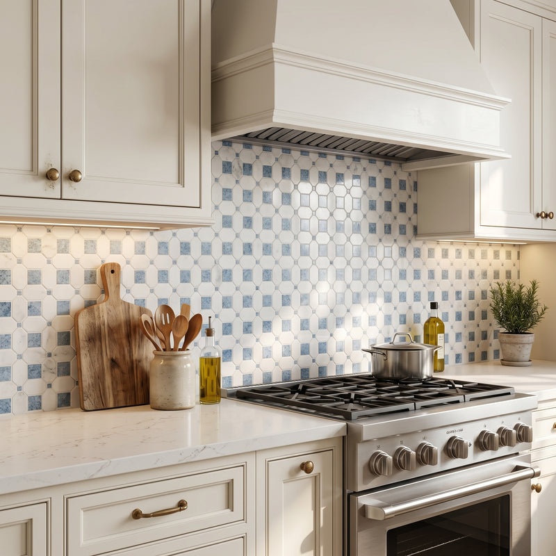 A kitchen scene with cream cabinetry, a white marble countertop, a stainless steel gas range, and a blue-and-white patterned mosaic tile backsplash. A wooden cutting board, utensil holder, olive oil bottle, and small potted plant sit on the counter.