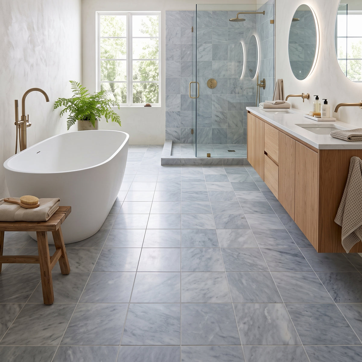 A bright, modern bathroom with large light gray marble-look floor tiles, a white freestanding bathtub, a wood vanity with white sinks, and a glass shower enclosure in the background.