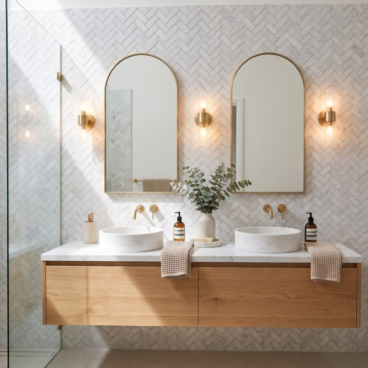 A modern double-sink bathroom vanity with two arched mirrors, gold wall sconces, a floating light wood cabinet, white vessel sinks, and a white herringbone tile wall.