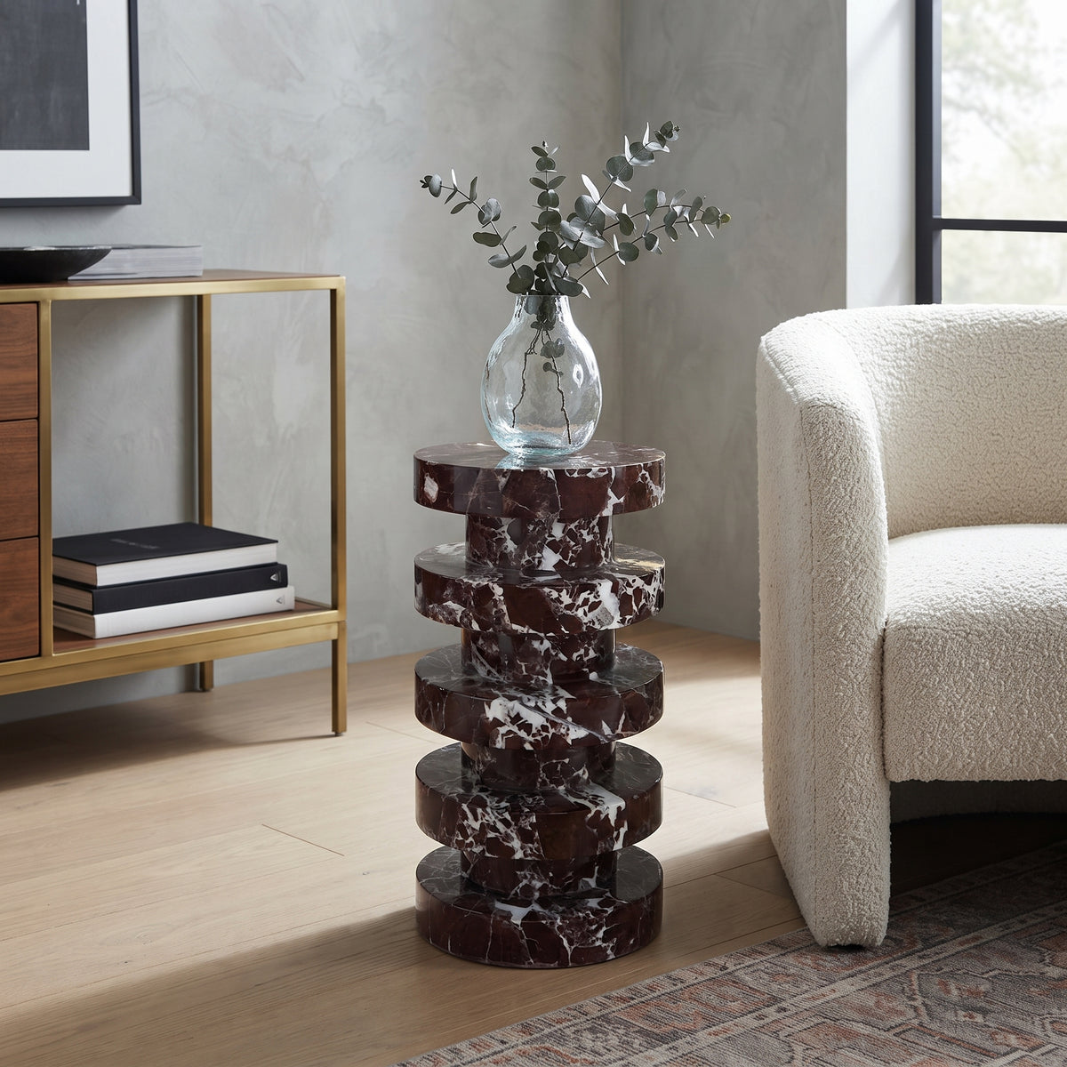 A modern spiral stack side table made of dark reddish-brown and white marble, styled in a living room next to a cream boucle armchair and a gold console table.