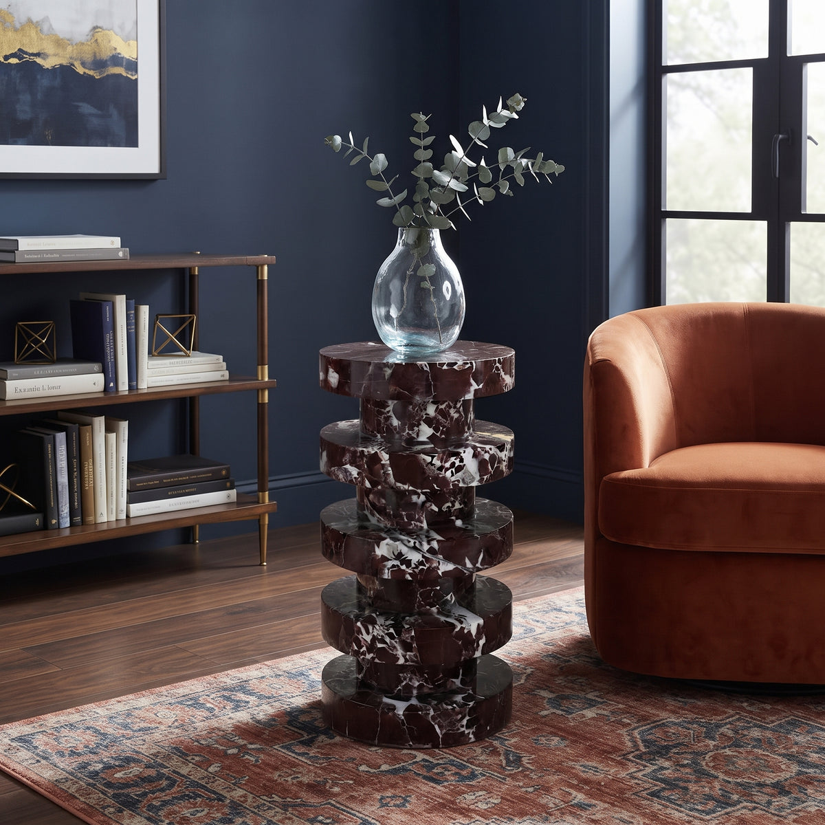 A round stacked side table made of dark reddish-brown and white marble, styled in a living room with a glass vase of eucalyptus on top, next to an orange armchair and a bookshelf.