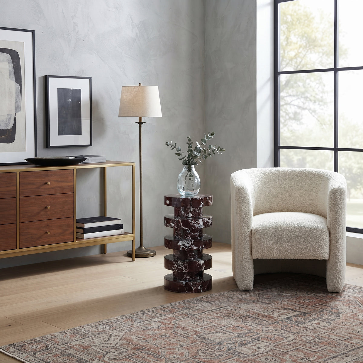 A modern living room scene featuring a cream bouclé armchair next to a dark red stacked marble side table holding a glass vase with greenery. A gold-framed wood console cabinet, a floor lamp, and framed wall art are arranged in a styled interior with a gray textured wall, light wood floors, and a patterned rug.