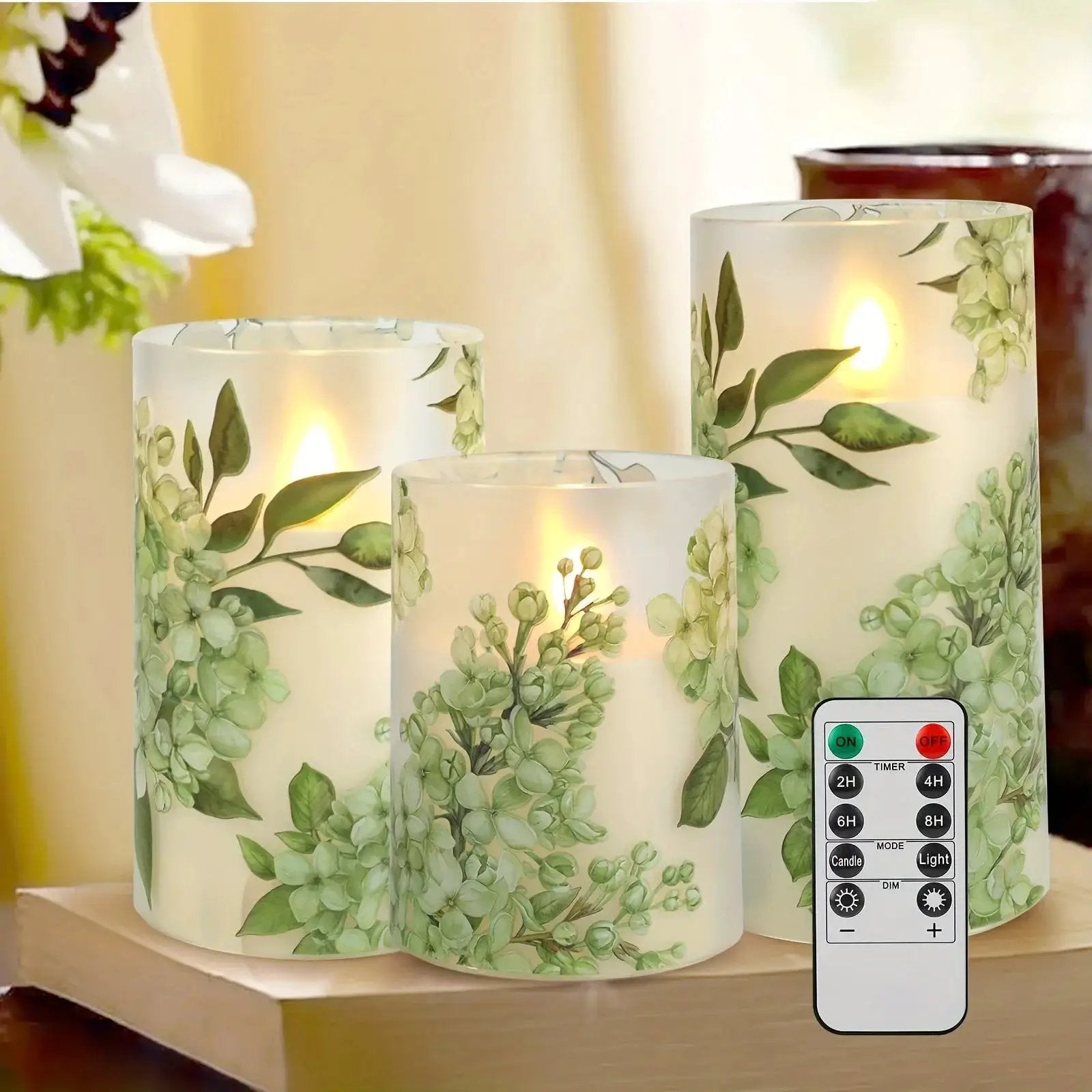 A set of 3 flameless candles, flickering LED pillar candles, with a 10-button remote control. They feature a green floral theme, are suitable for spring and summer, and can be used on the dining tables at wedding venues by House Of Avana