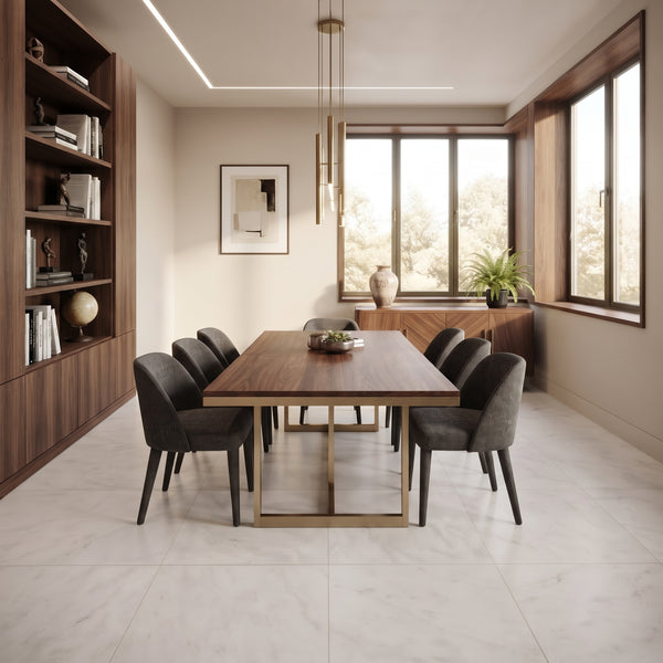 A modern dining room featuring a rectangular wooden dining table with a gold metal base, surrounded by six dark gray upholstered chairs. The room has light beige walls, a large built-in wooden bookshelf on the left, and large windows with a sideboard and potted plants on the right.