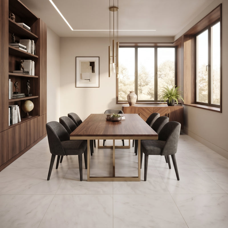 A modern dining room featuring a rectangular wooden dining table with a gold metal base, surrounded by six dark gray upholstered chairs. The room has light beige walls, a large built-in wooden bookshelf on the left, and large windows with a sideboard and potted plants on the right.