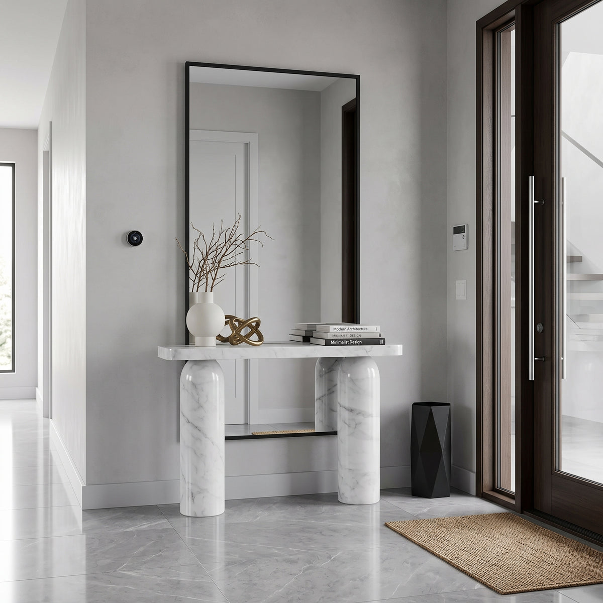 A modern console table with a white marble top and cylindrical marble pedestal legs, positioned beneath a tall rectangular black-framed mirror in a minimalist entryway. Decorative items including a white vase with dried branches, a gold sculpture, and a stack of books sit on the tabletop.