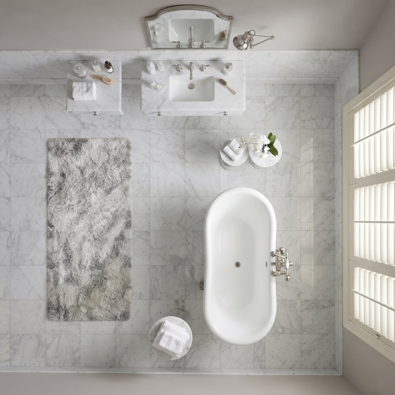 Overhead view of a modern bathroom with a white freestanding bathtub, marble-look tiled walls, a gray marbled bath rug, a white sink vanity with mirror, and a window with white shutters.
