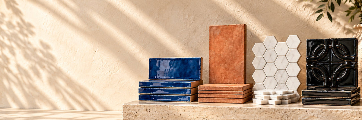 Stacks of glazed blue ceramic, terracotta, white marble hexagon and black relief tiles on a travertine ledge in afternoon sun