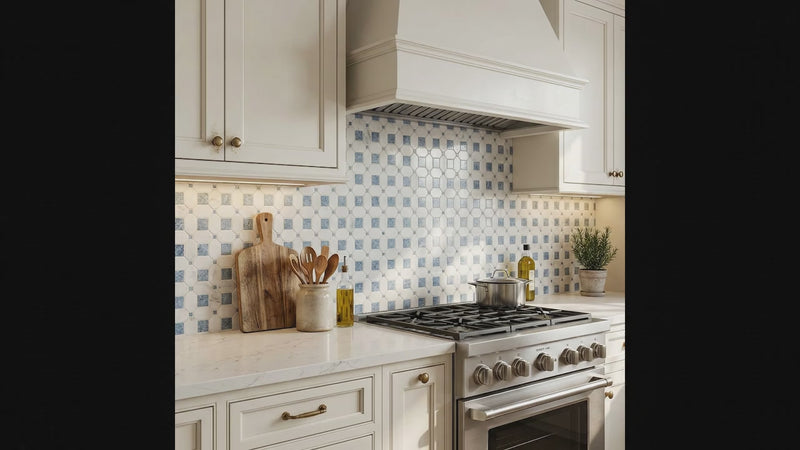 A kitchen scene with cream cabinetry, a white marble countertop, a stainless steel gas range, and a blue-and-white patterned mosaic tile backsplash. A wooden cutting board, utensil holder, olive oil bottle, and small potted plant sit on the counter.