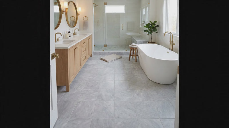 A bright, modern bathroom featuring large light gray marble-look floor tiles, a white freestanding bathtub with brass fixtures, a double-sink vanity with light wood cabinets, and a glass shower enclosure.