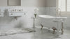 a modern bathroom with a white freestanding bathtub, marble-look tiled walls, a gray marbled bath rug, a white sink vanity with mirror, and a window with white shutters.