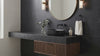 Modern floating bathroom vanity with a black stone countertop and sink, dark wood cabinet, round wall mirror, and matching sconces against a black tiled wall.