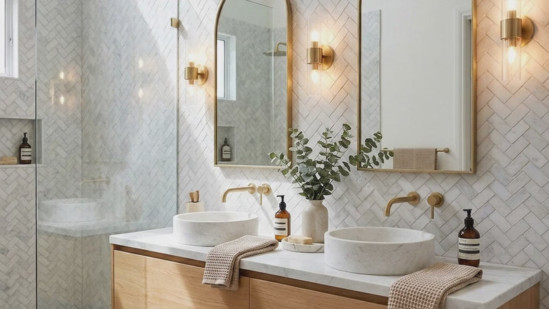 Modern bathroom vanity with two vessel sinks, a floating wood cabinet, white marble countertop, two arched mirrors with gold frames, and warm wall sconces. A glass shower enclosure is visible on the left, and the wall behind features light herringbone tile.