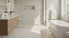 A bright, modern bathroom featuring light beige stone-look floor and wall tiles, a white freestanding bathtub with gold fixtures, a double-sink vanity, and a walk-in shower area.