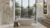 A luxury modern bathroom featuring a white freestanding oval bathtub, a glass shower enclosure, a floating vanity with a marble countertop, and large floor-to-ceiling windows overlooking a tropical garden.