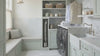 A bright, modern laundry room with sage green cabinetry, marble countertops and backsplash, a built-in window seat with pillows, a brass pendant light, and a patterned rug on a marble tile floor.