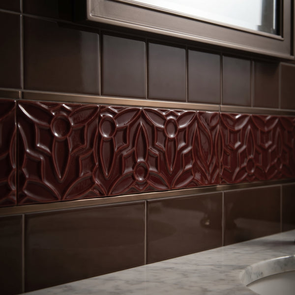 Decorative bathroom wall tile featuring a central raised relief band in deep burgundy-brown with a repeating floral pattern, flanked by glossy dark brown subway tiles and a marble countertop.