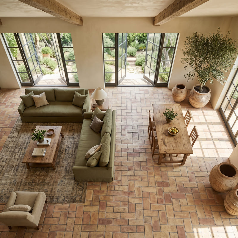 An overhead view of a rustic, high-ceilinged living room with olive green sofas, a wooden dining table, terracotta brick flooring, and large windows looking out to a garden.