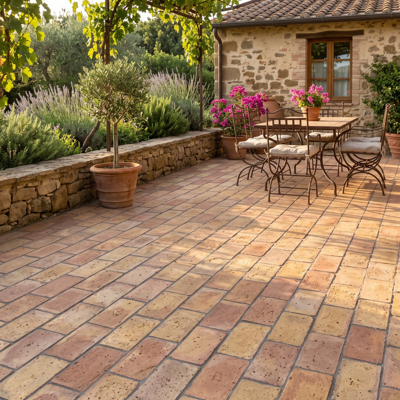 Terracotta paver tiles arranged on an outdoor patio in front of a rustic stone house, with a dining set, potted plants, and greenery visible.