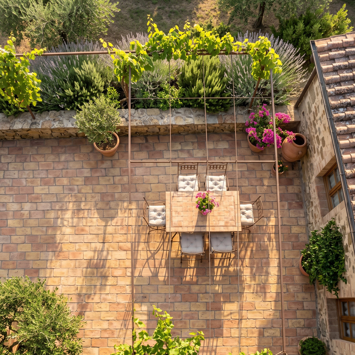 An overhead view of a rustic stone patio featuring a wooden dining table with four cushioned chairs, a small flower arrangement, a metal trellis with climbing green vines, and potted plants against a brick wall.