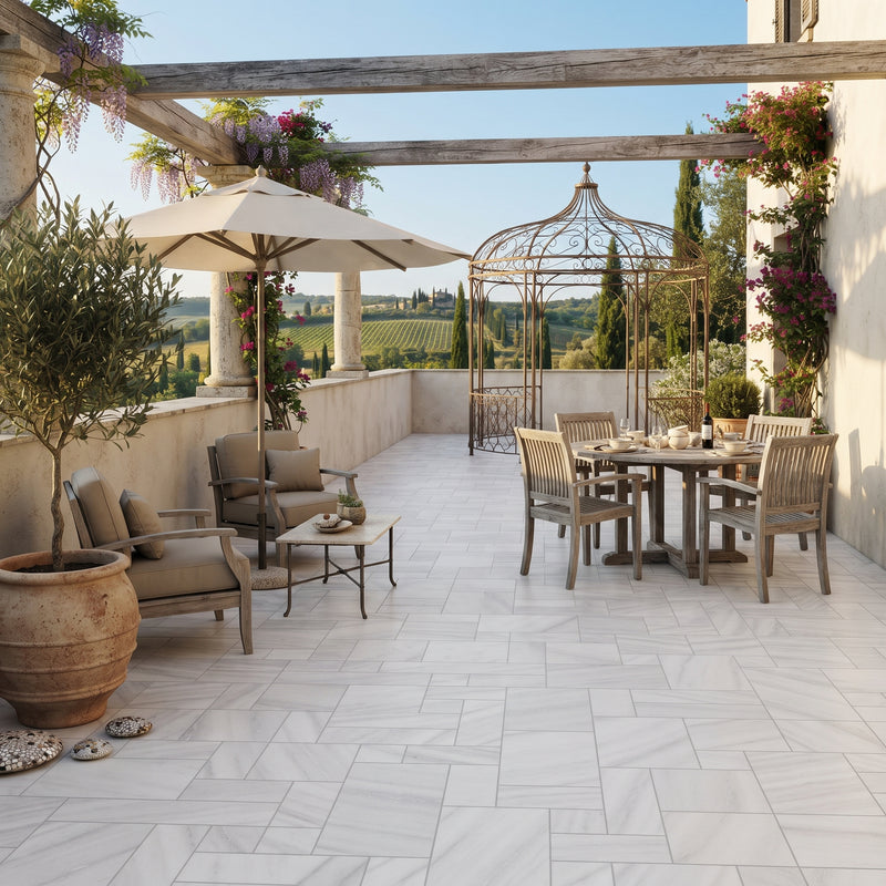 A luxury outdoor patio scene featuring light gray marble-look rectangular tiles, a dining set with woven chairs and a wooden table, a beige patio umbrella, a metal gazebo, and a large potted olive tree under a wooden pergola with climbing plants.