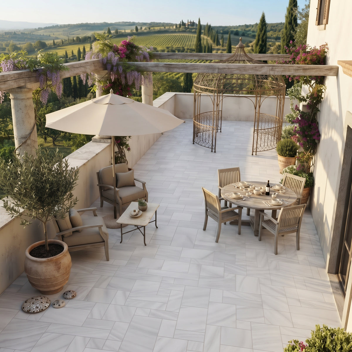 An outdoor patio with a light stone tile floor, furnished with a dining set, lounge chairs, a coffee table, and a large umbrella. The terrace is decorated with potted plants, a pergola with hanging flowers, and a scenic vineyard landscape in the background.