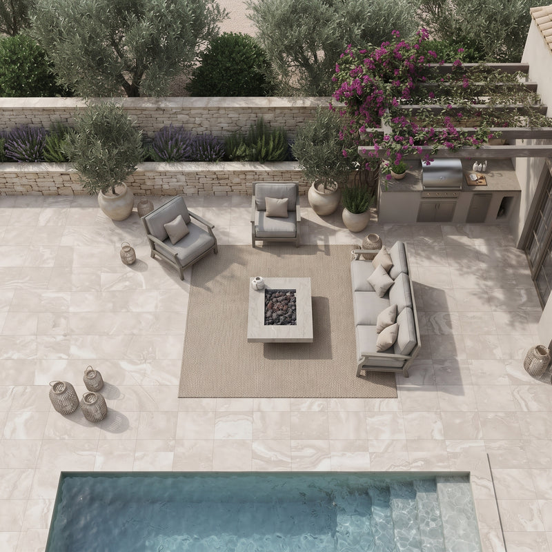 An overhead view of a luxury outdoor patio featuring a light beige stone tile floor, a built-in outdoor kitchen with a stainless steel grill, a rectangular fire pit table on a central beige rug, a gray three-seater sofa, two matching armchairs, and a swimming pool in the foreground.